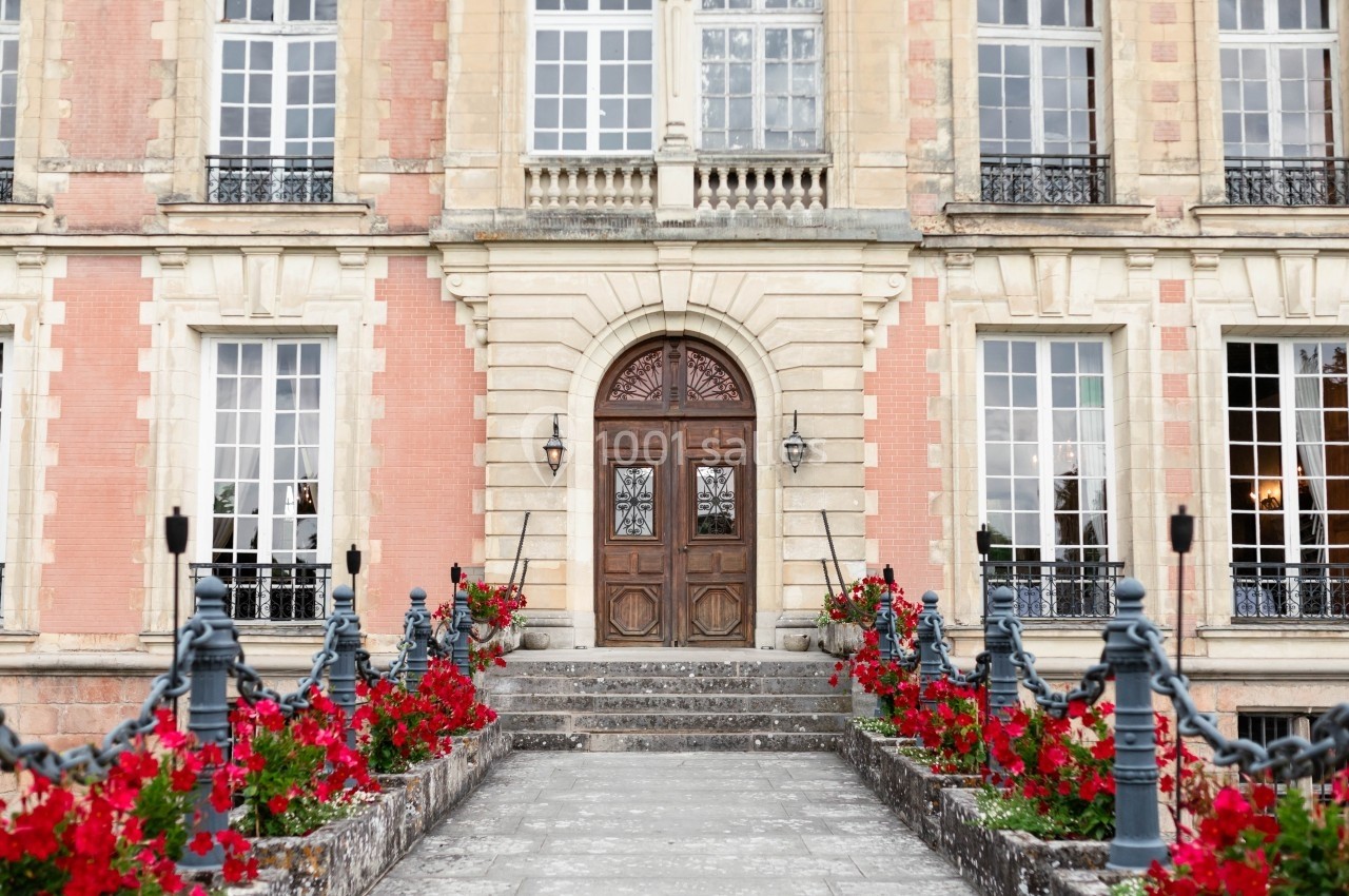 Façade d'un bâtiment ancien avec porte en bois sculpté, grandes fenêtres et allée bordée de fleurs rouges.