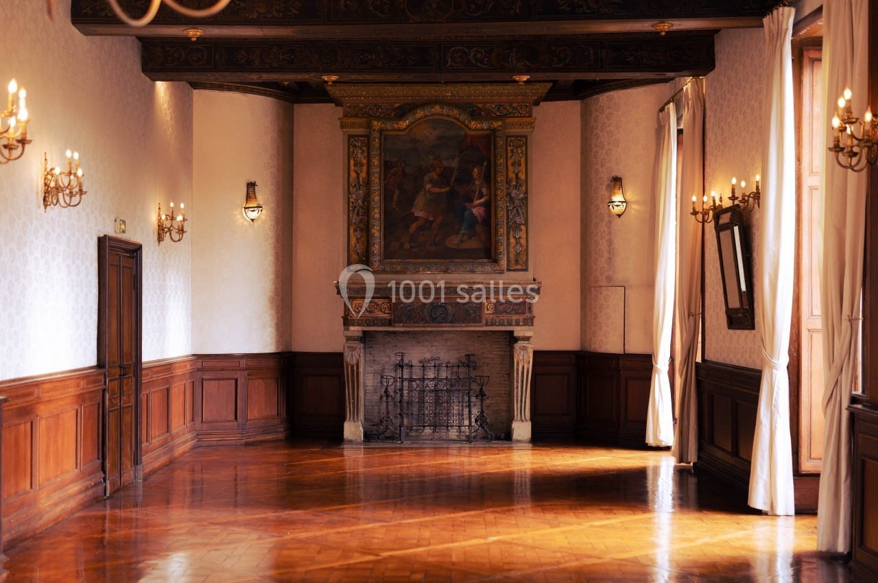 Salle ancienne avec parquet en bois, cheminée ornée d'une peinture, murs lambrissés et éclairage par appliques murales.