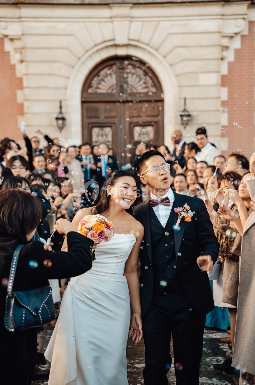 Un couple souriant sort d'un bâtiment après leur mariage, entouré d'invités et de bulles de savon.