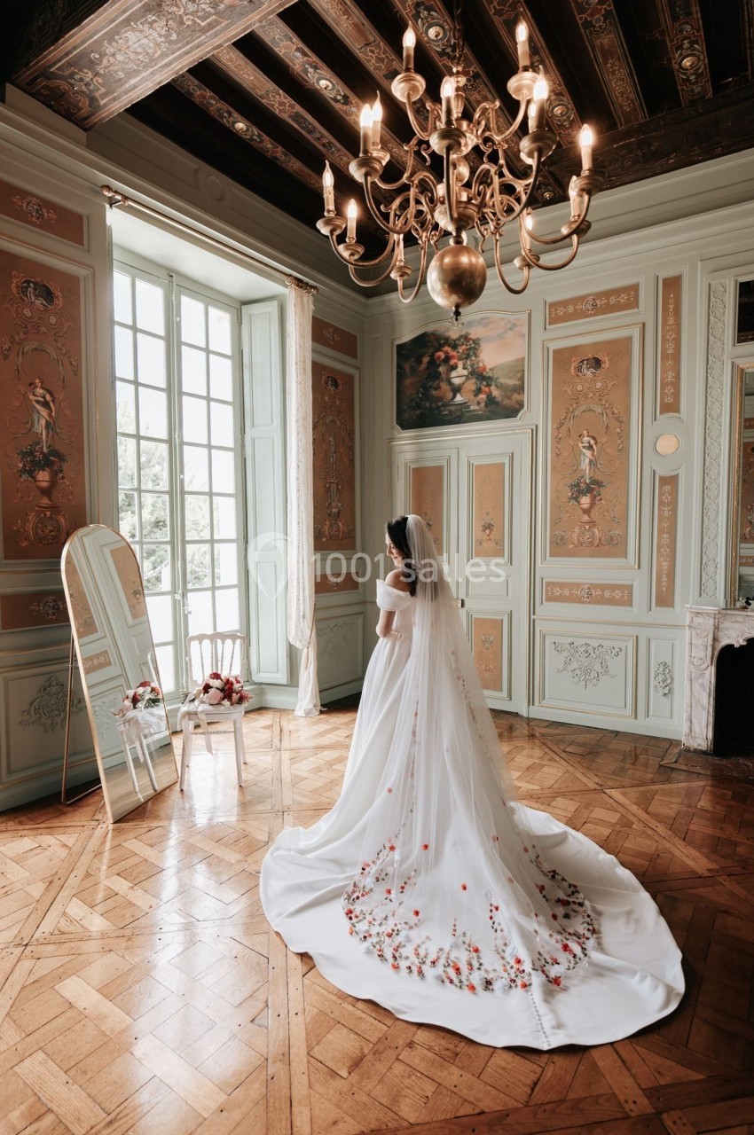 Une mariée en robe blanche avec une longue traîne brodée de fleurs, debout dans une pièce élégante au décor classique.