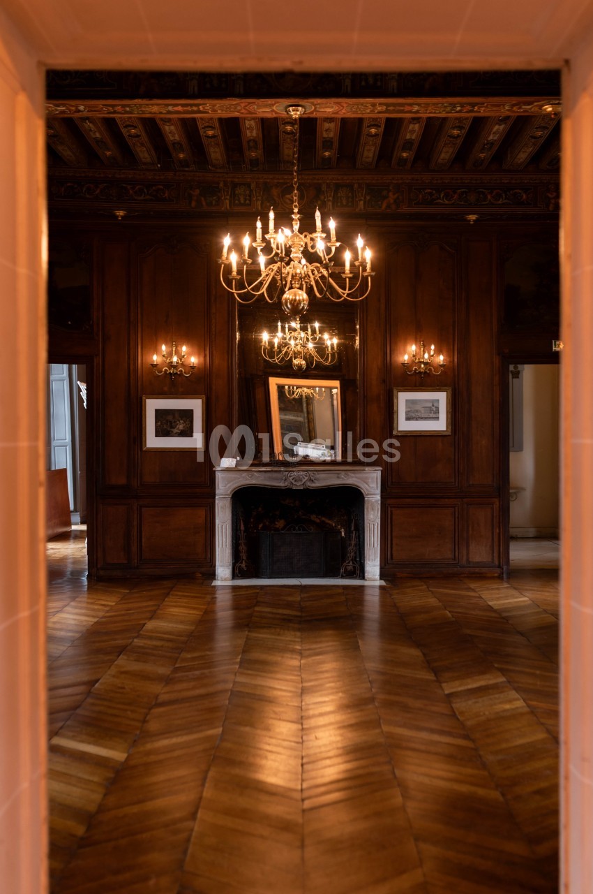 Salle aux boiseries brunes, cheminée centrale, lustres dorés et parquet en chevrons.