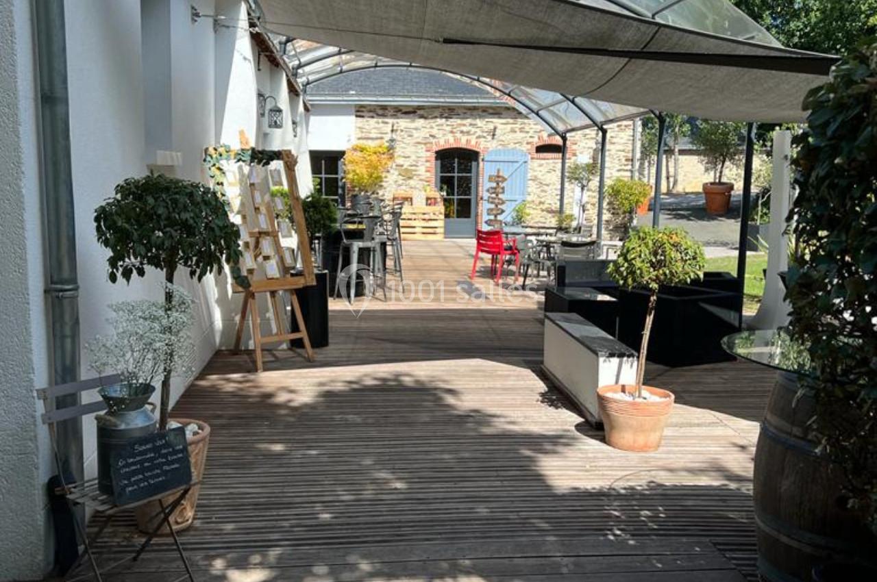Terrasse en bois avec tables, chaises, plantes en pot et chevalets, partiellement couverte par une structure transparente.
