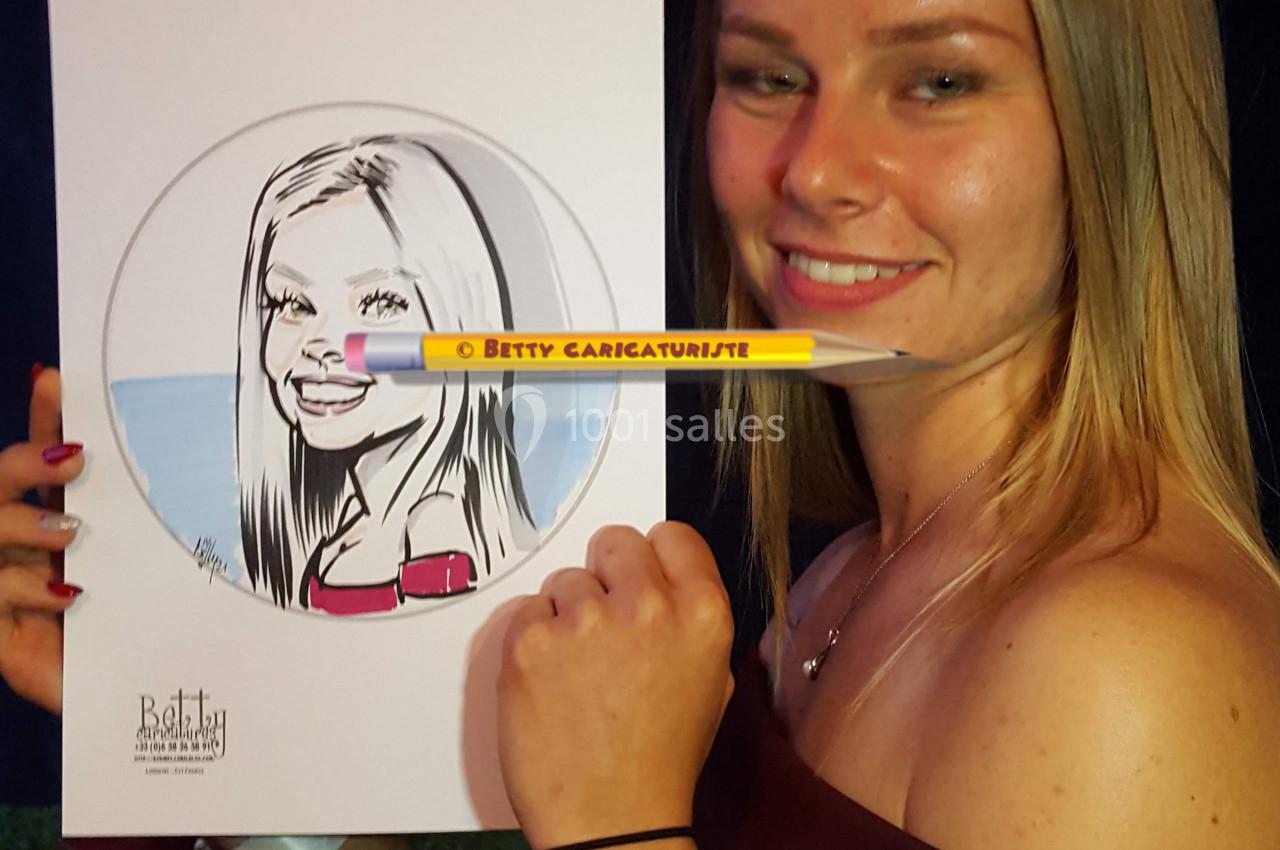 Une femme souriante tient un portrait caricatural d'elle-même, réalisé à la main, dans un cadre extérieur.
