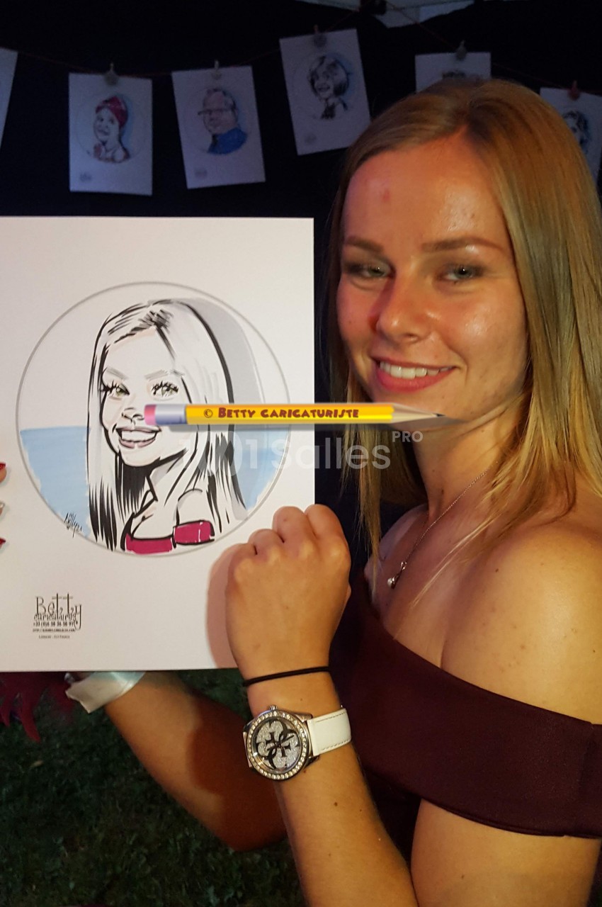 Une femme souriante tient un portrait caricatural d'elle-même, réalisé à la main, dans un cadre extérieur.