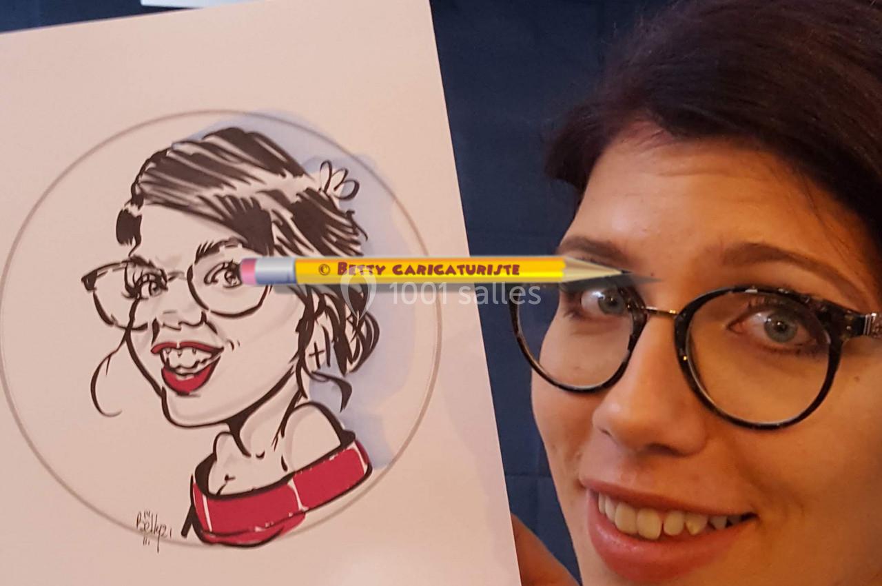 Femme souriante tenant une caricature d'elle-même, réalisée sur papier, avec d'autres dessins exposés en arrière-plan.