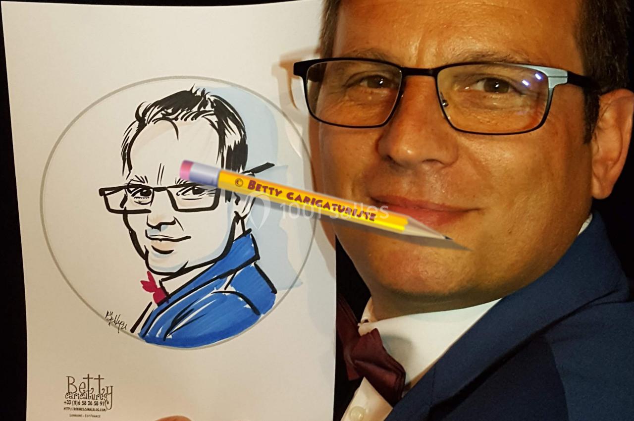 Homme en costume tenant un dessin caricatural de lui-même, avec des portraits accrochés en arrière-plan.