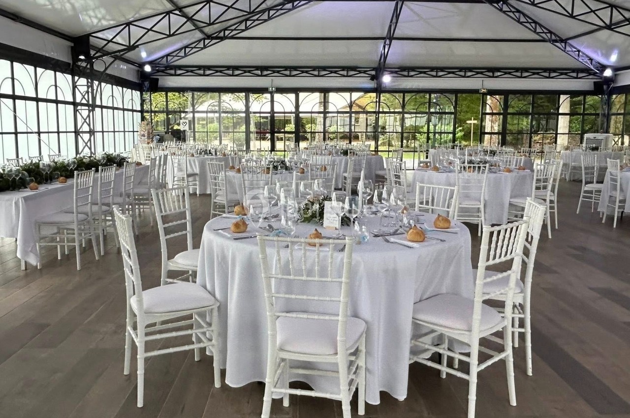 Salle de réception lumineuse avec tables rondes dressées, nappes blanches et chaises en bois clair.