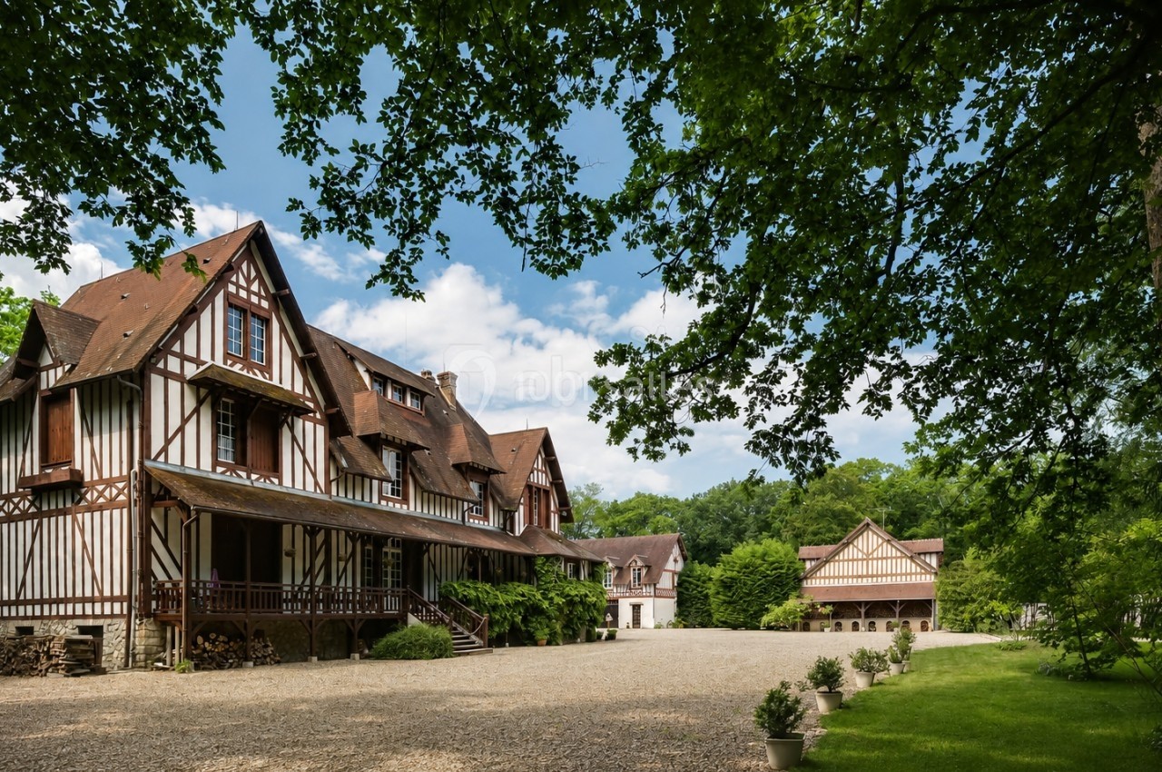 Grand bâtiment à colombages entouré d'arbres, avec une cour en gravier et un jardin verdoyant.
