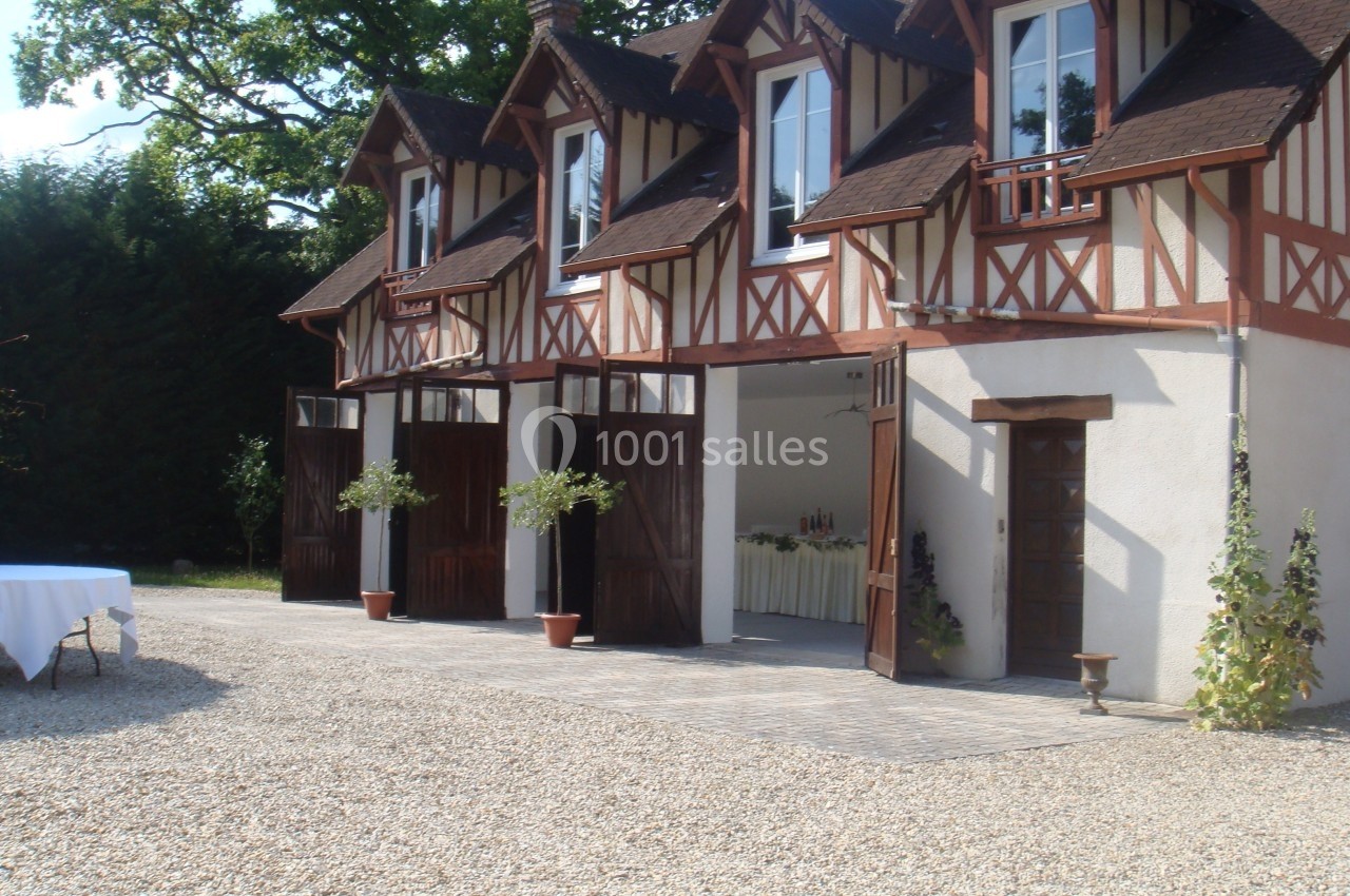 Façade d'un bâtiment à colombages avec portes ouvertes, donnant sur une cour en gravier et des arbres en arrière-plan.