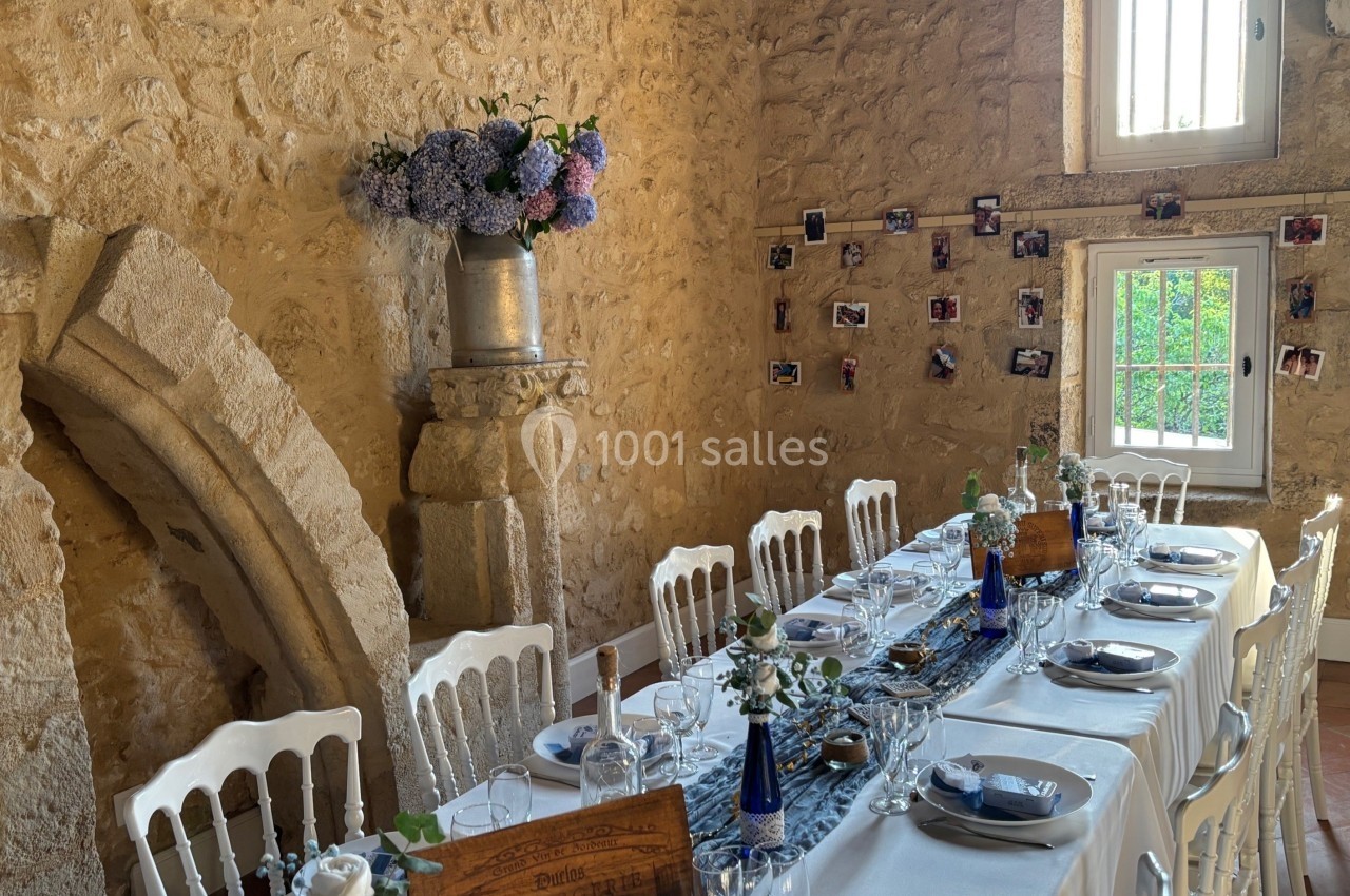 Table décorée pour un repas dans une salle en pierre, avec fleurs, photos accrochées au mur et lumière naturelle.