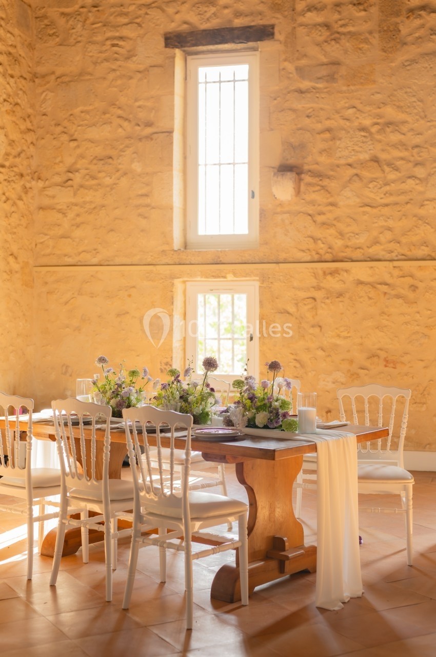 Table en bois décorée de fleurs et entourée de chaises blanches dans une salle en pierre avec lumière naturelle.