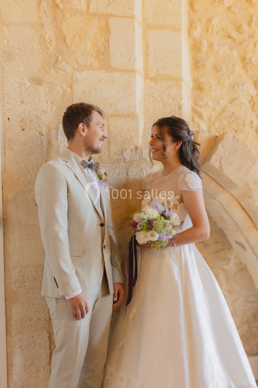 Un couple en tenue de mariage se tient debout près d'un mur en pierre, échangeant un regard souriant.