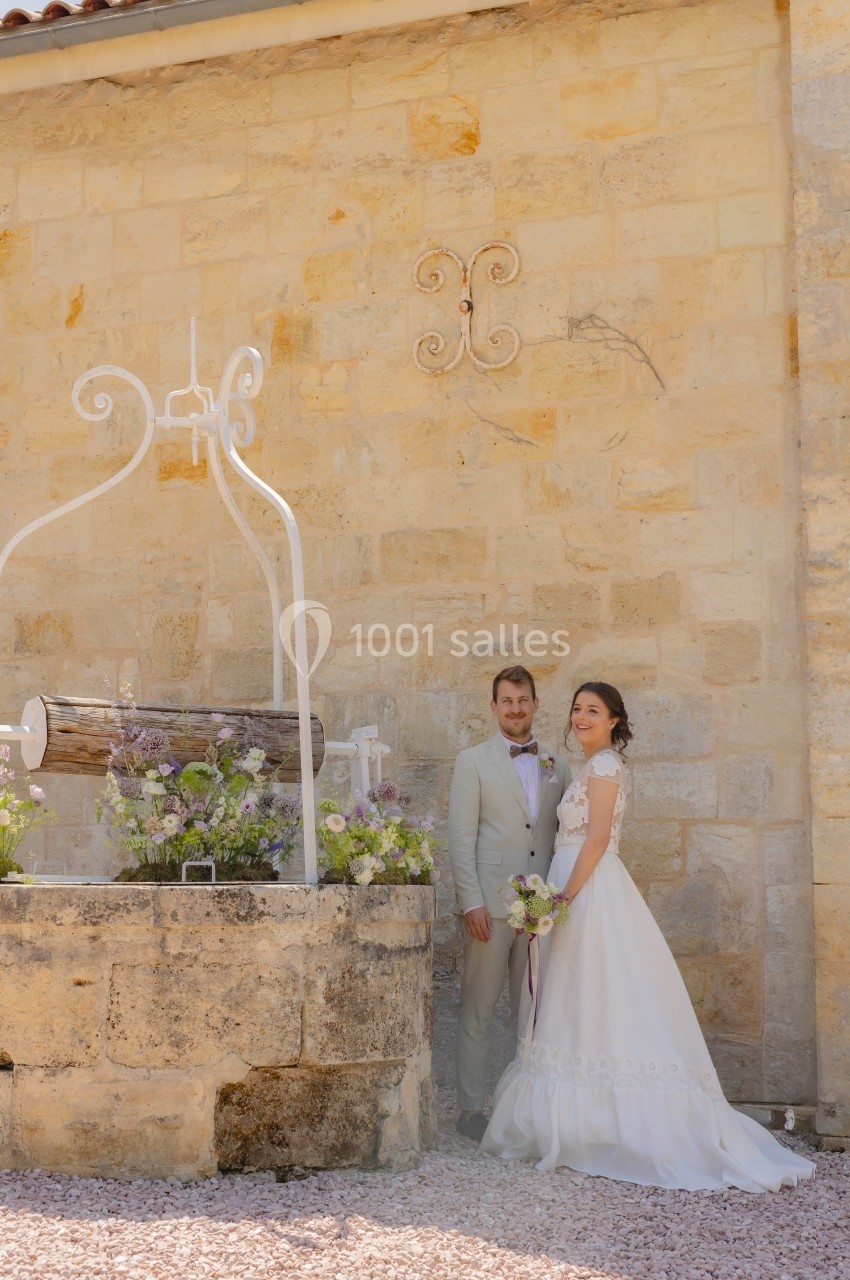 Un couple en tenue de mariage pose près d'un puits décoré de fleurs devant un mur en pierre claire.