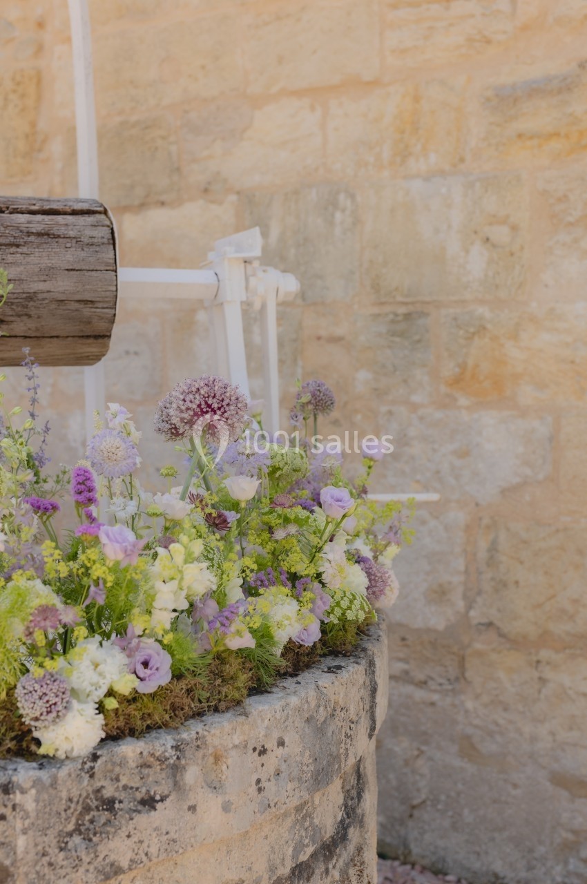 Fleurs violettes et blanches disposées sur un puits en pierre devant un mur beige en pierre.