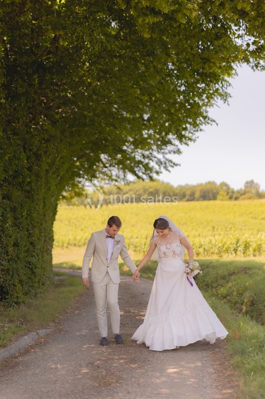 Un couple de mariés marche main dans la main sur un chemin bordé d'arbres, avec un champ en arrière-plan.