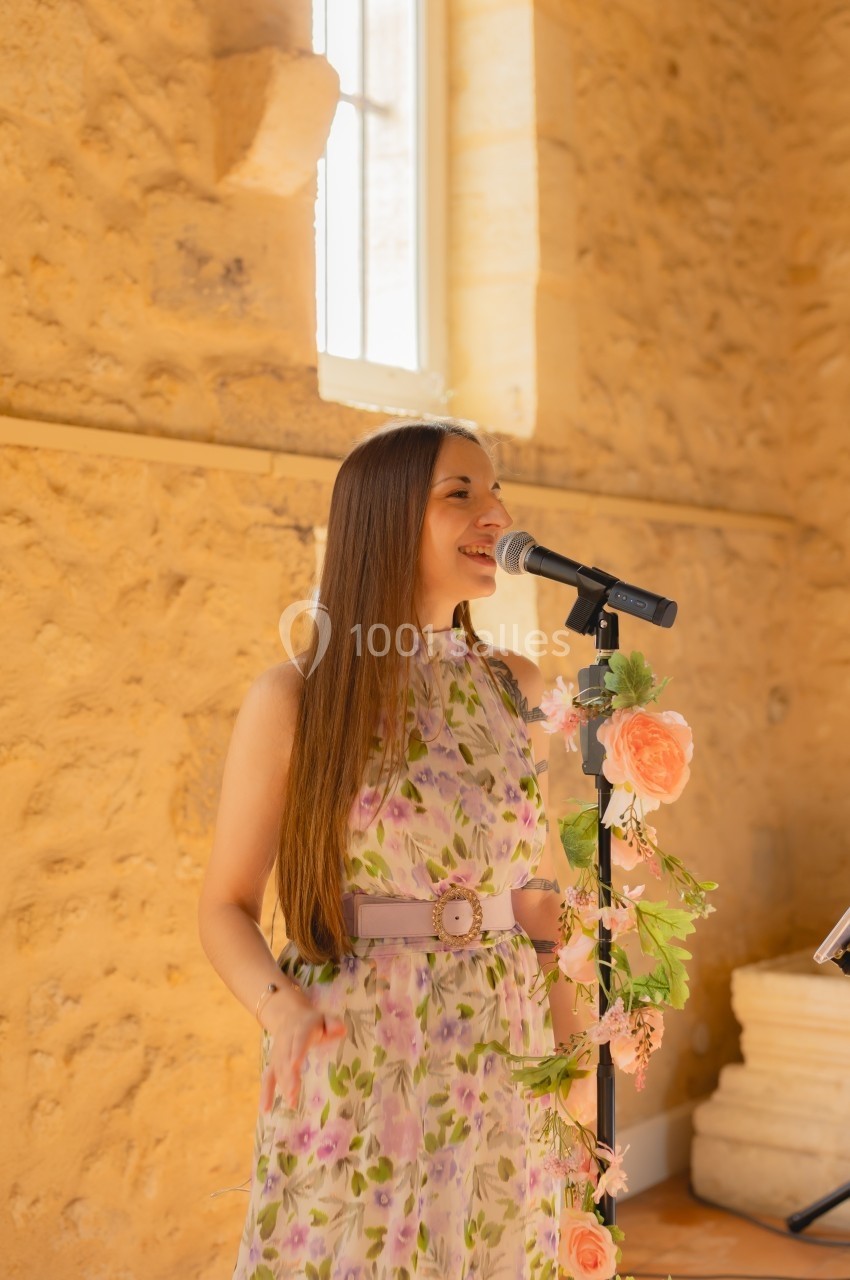 Une femme chante au micro dans une salle en pierre éclairée par la lumière naturelle, décorée de fleurs.