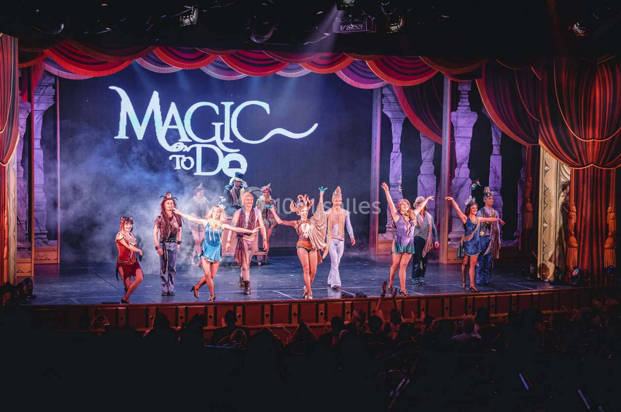 Spectacle sur scène avec des artistes costumés dansant devant un décor lumineux et un rideau rouge.