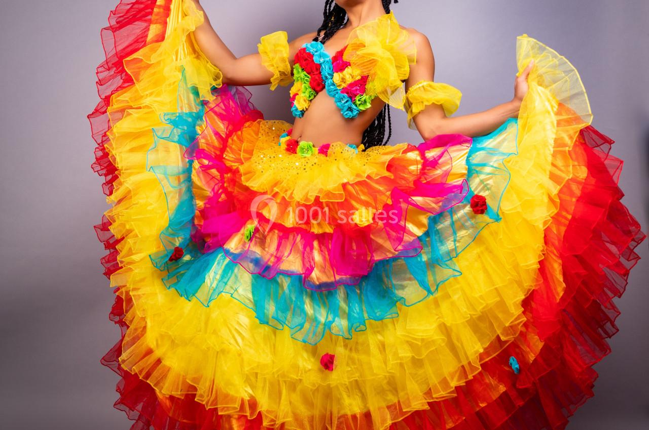 Femme portant une robe colorée à volants jaunes, rouges et bleus, posant devant un fond gris.