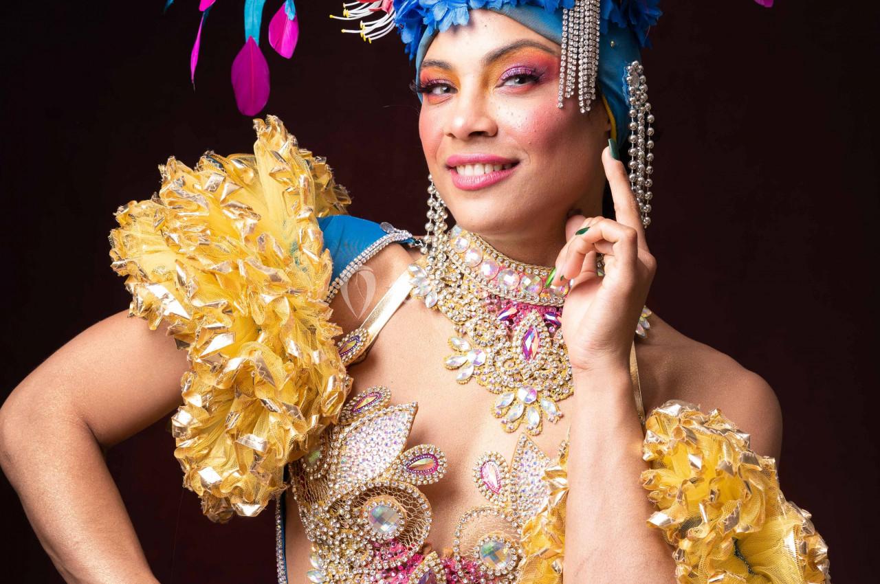 Personne en costume coloré avec plumes, perles et fleurs, posant devant un fond sombre.