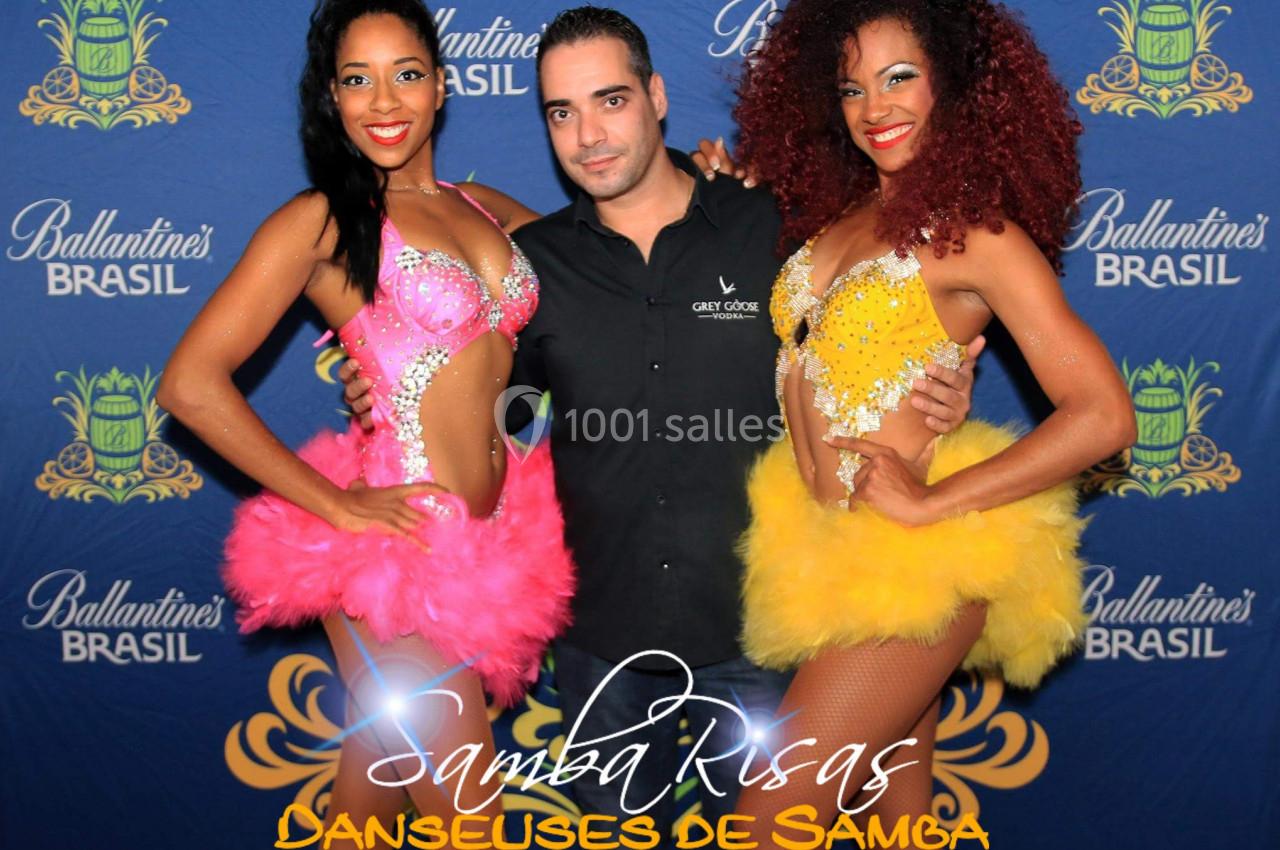 Un homme pose avec deux danseuses en costumes colorés devant un fond promotionnel ’Ballantine's Brasil’.