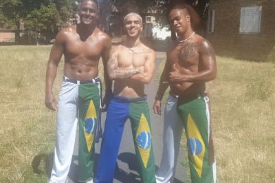 Miniature Sambarisas #6 Trois hommes souriants en tenue de capoeira avec des drapeaux brésiliens, debout dans un parc ensoleillé.