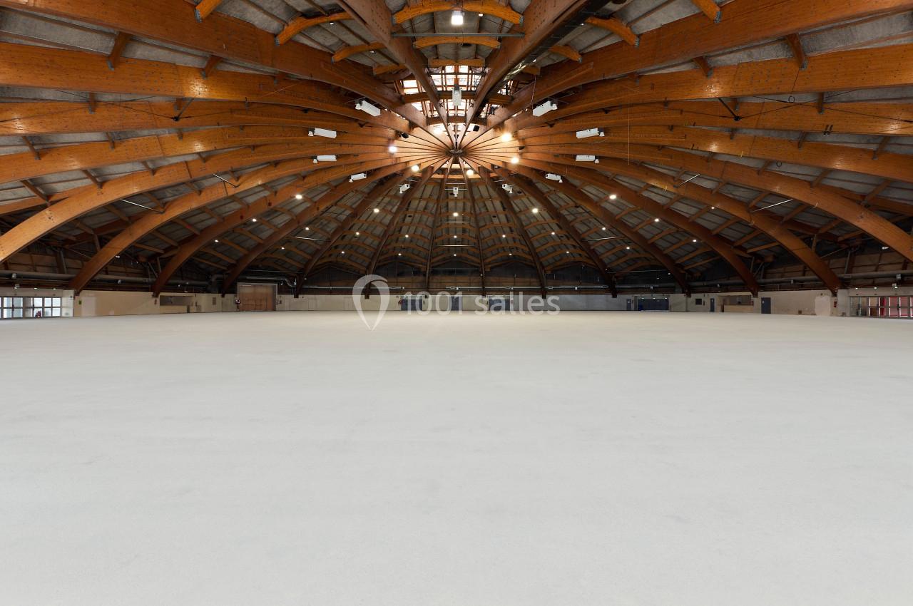 Vue intérieure d'un vaste hall avec une structure en bois courbée et un sol clair, éclairé par des ouvertures au plafond.