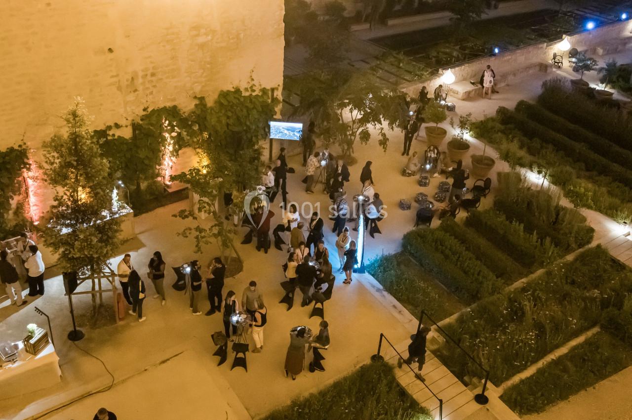Vue aérienne d'une cour éclairée en soirée, avec des invités rassemblés autour de tables dans un jardin structuré.