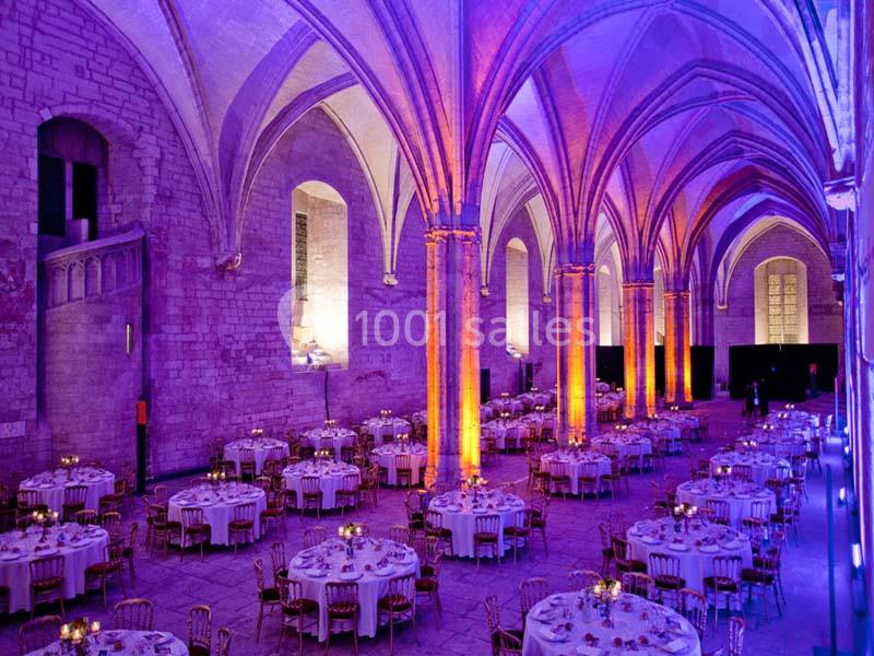 Salle voûtée éclairée en violet et orange, aménagée avec des tables rondes dressées pour un événement.