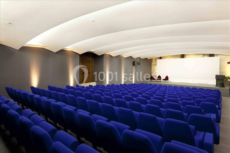 Salle de conférence avec rangées de sièges bleus, écran blanc en fond et éclairage discret au plafond.