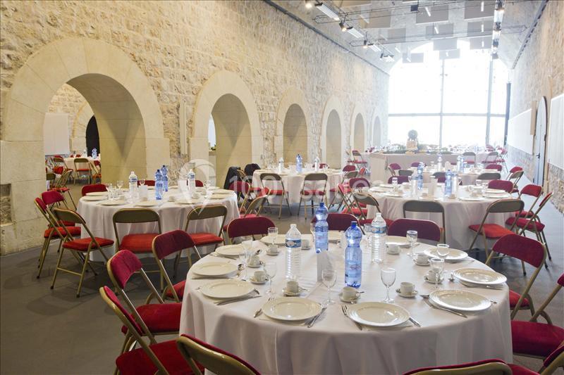 Salle en pierre avec des tables rondes dressées, chaises rouges et bouteilles d'eau, prête pour un événement.