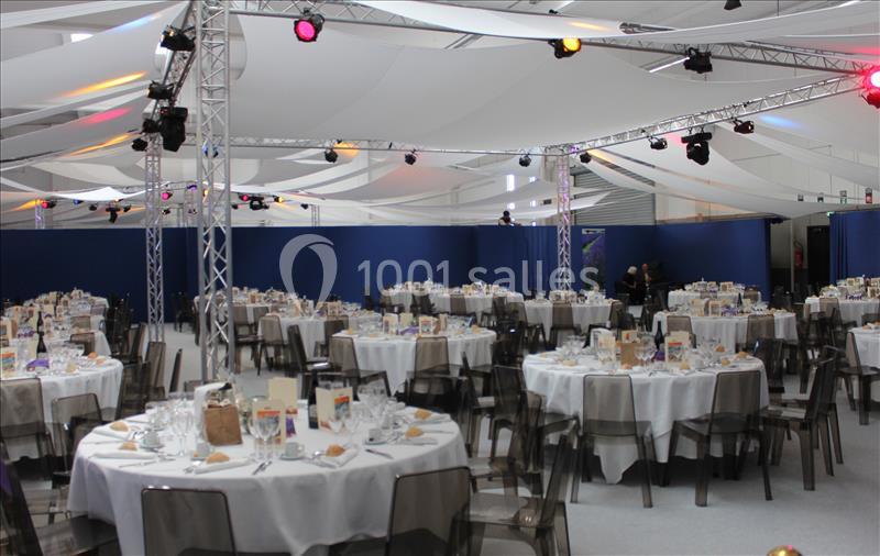 Salle de réception avec tables rondes dressées, nappes blanches, chaises transparentes et éclairage suspendu.
