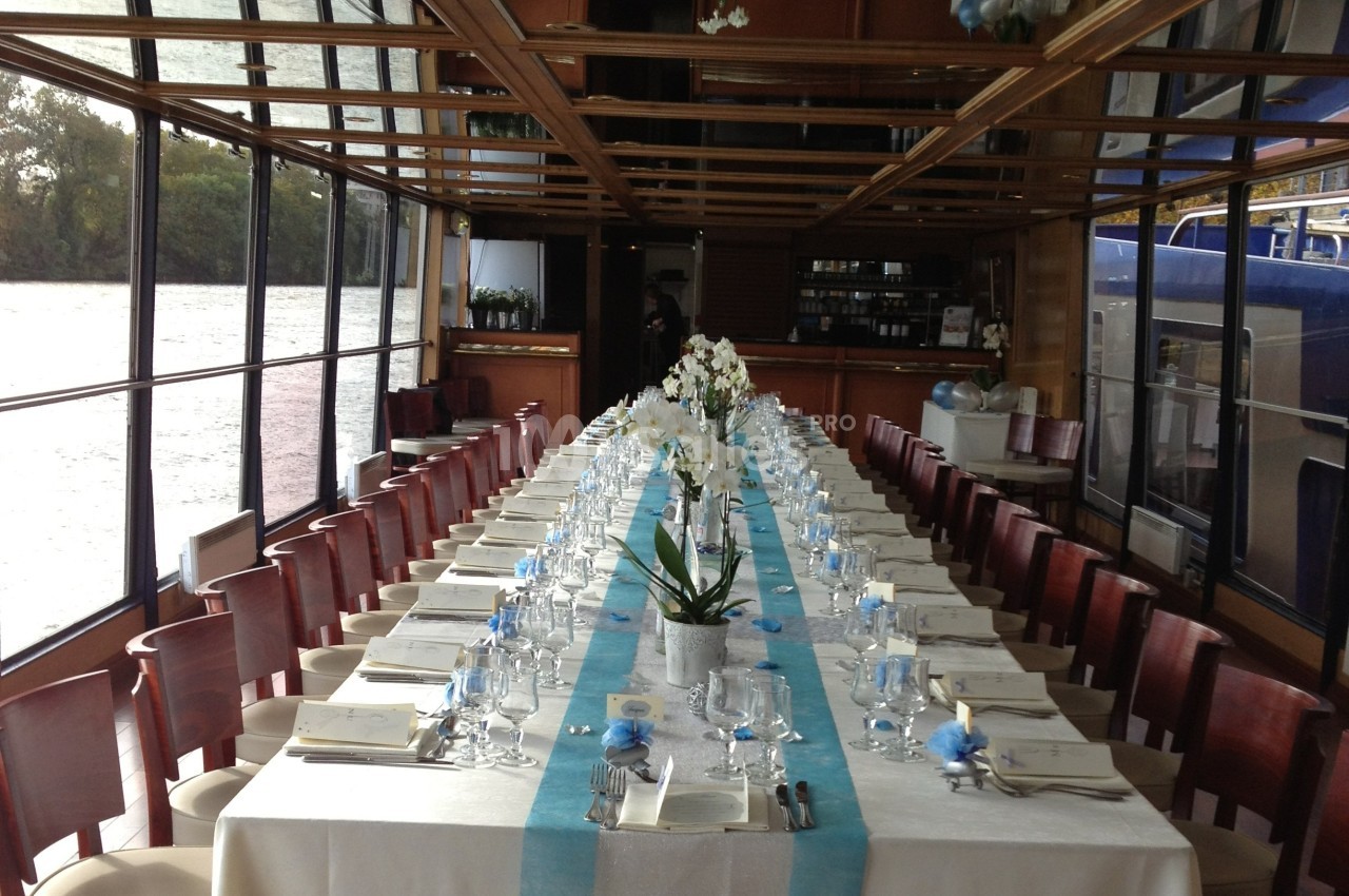 Salle de réception sur un bateau avec une longue table dressée, ornée de fleurs blanches et décorations bleues.