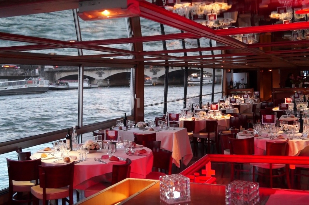Salle de restaurant avec tables dressées, baies vitrées donnant sur une rivière et un pont visible à l'extérieur.