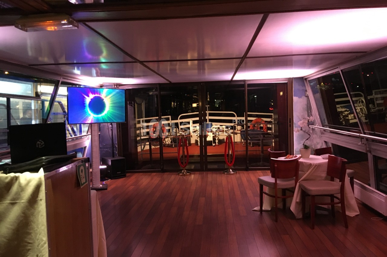 Intérieur d'un bateau avec parquet en bois, écran lumineux, tables dressées et vue sur le pont extérieur éclairé.