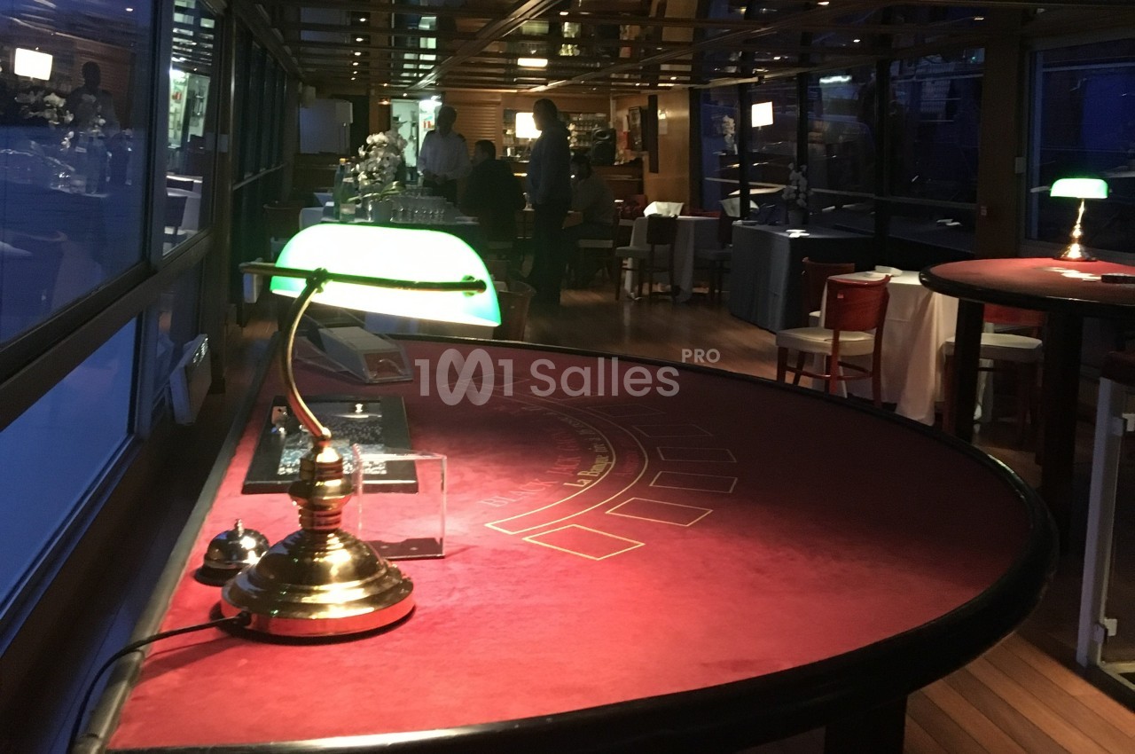 Table de jeu rouge éclairée par une lampe verte dans une salle avec des baies vitrées et un bar en arrière-plan.