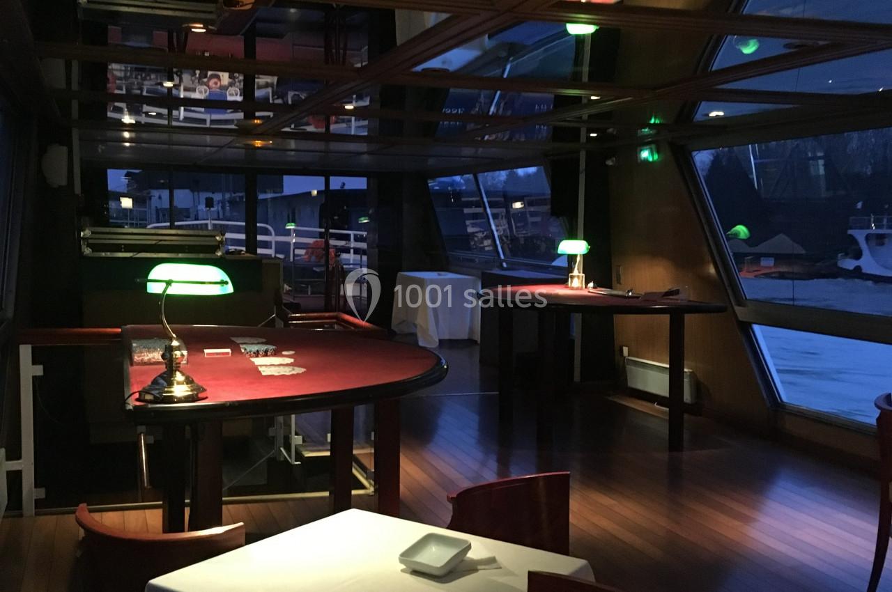 Salle avec tables de jeux, lampes vertes et grandes baies vitrées donnant sur un extérieur sombre.