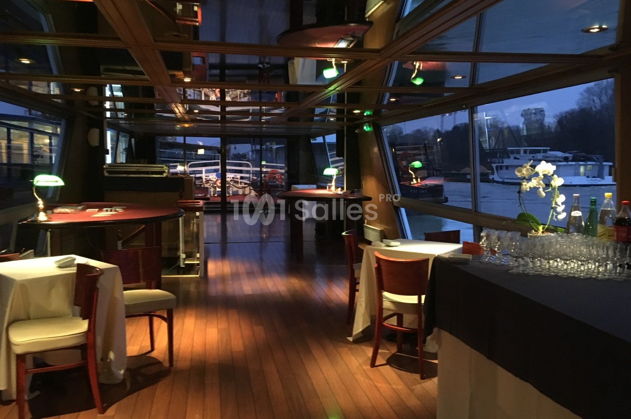 Salle de restaurant élégante avec parquet en bois, tables dressées et vue sur un quai à travers de grandes baies vitrées.
