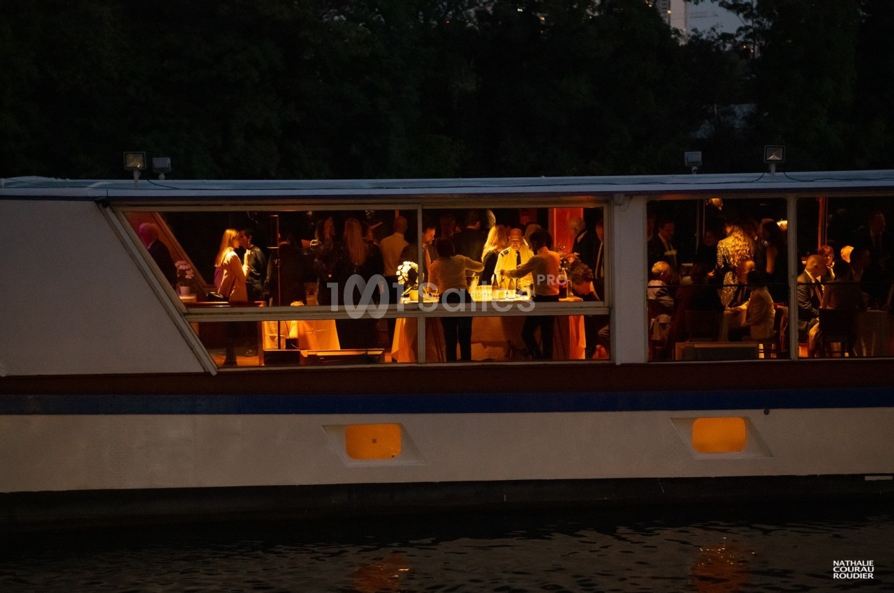 Des personnes participent à une réception éclairée à l'intérieur d'un bateau amarré, en soirée.