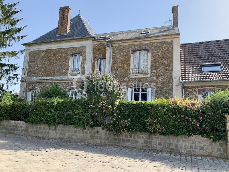 Façade d'une maison en pierre avec volets blancs, entourée de haies fleuries et donnant sur une rue pavée.