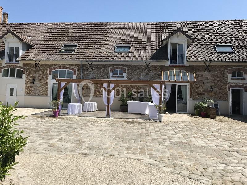 Façade d'une grande bâtisse en pierre avec toit en tuiles, tables décorées et pergola en bois sur une cour pavée.
