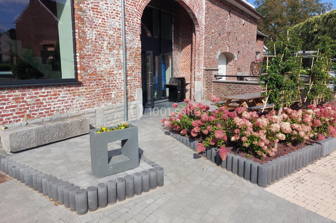 Entrée d'un bâtiment en briques rouges avec parterre de fleurs, pavés au sol et mobilier extérieur en bois.