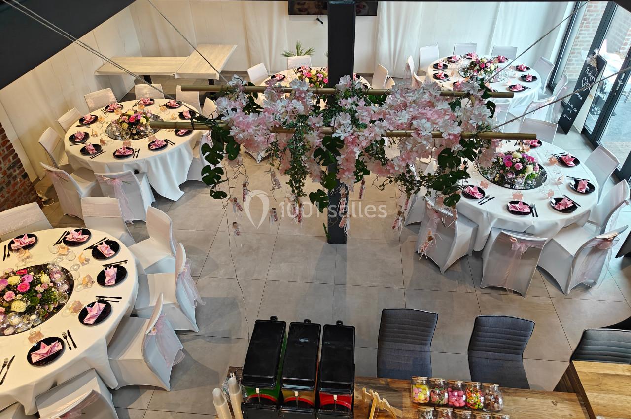 Salle décorée pour un événement avec tables rondes dressées, chaises blanches et centre de table fleuri suspendu.