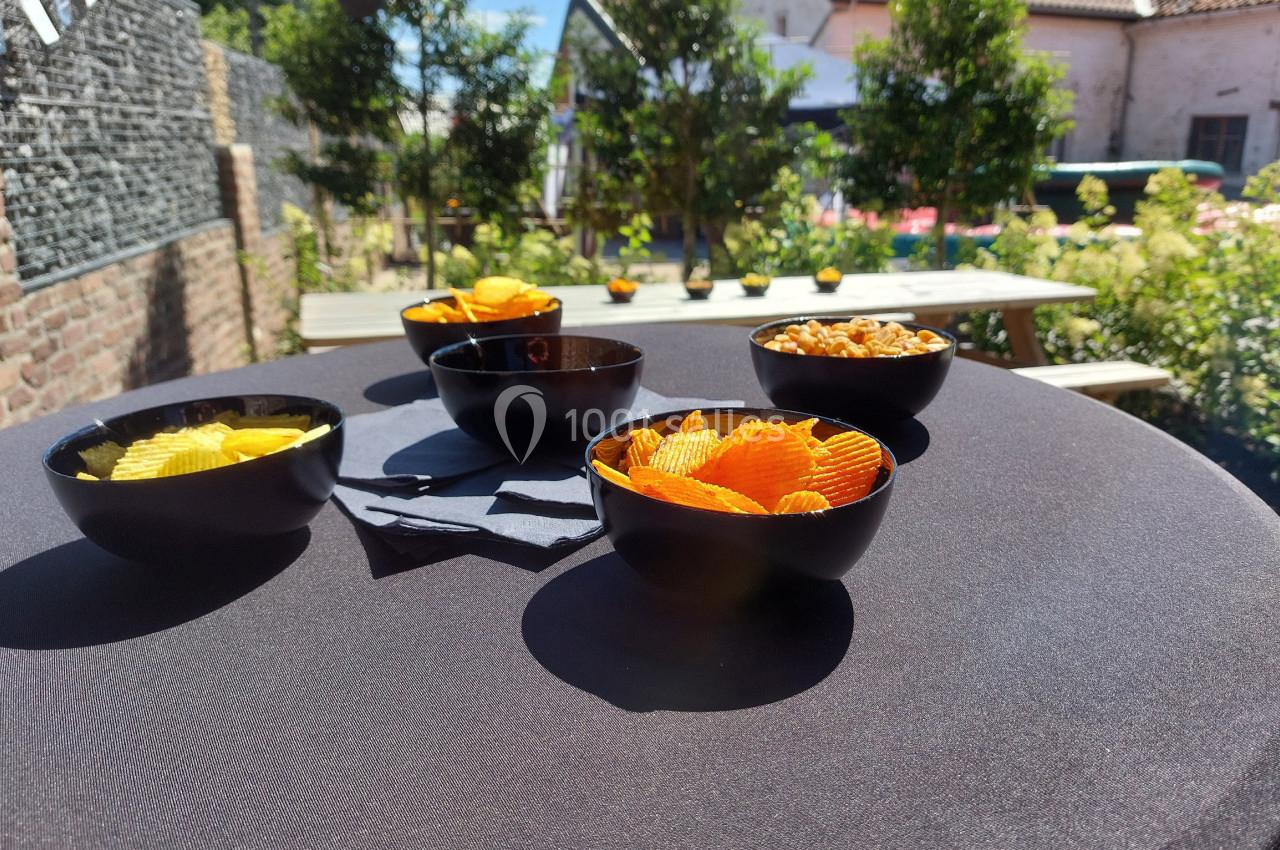 Quatre bols noirs contenant des chips et des snacks sont posés sur une table ronde en extérieur, sous un ciel ensoleillé.