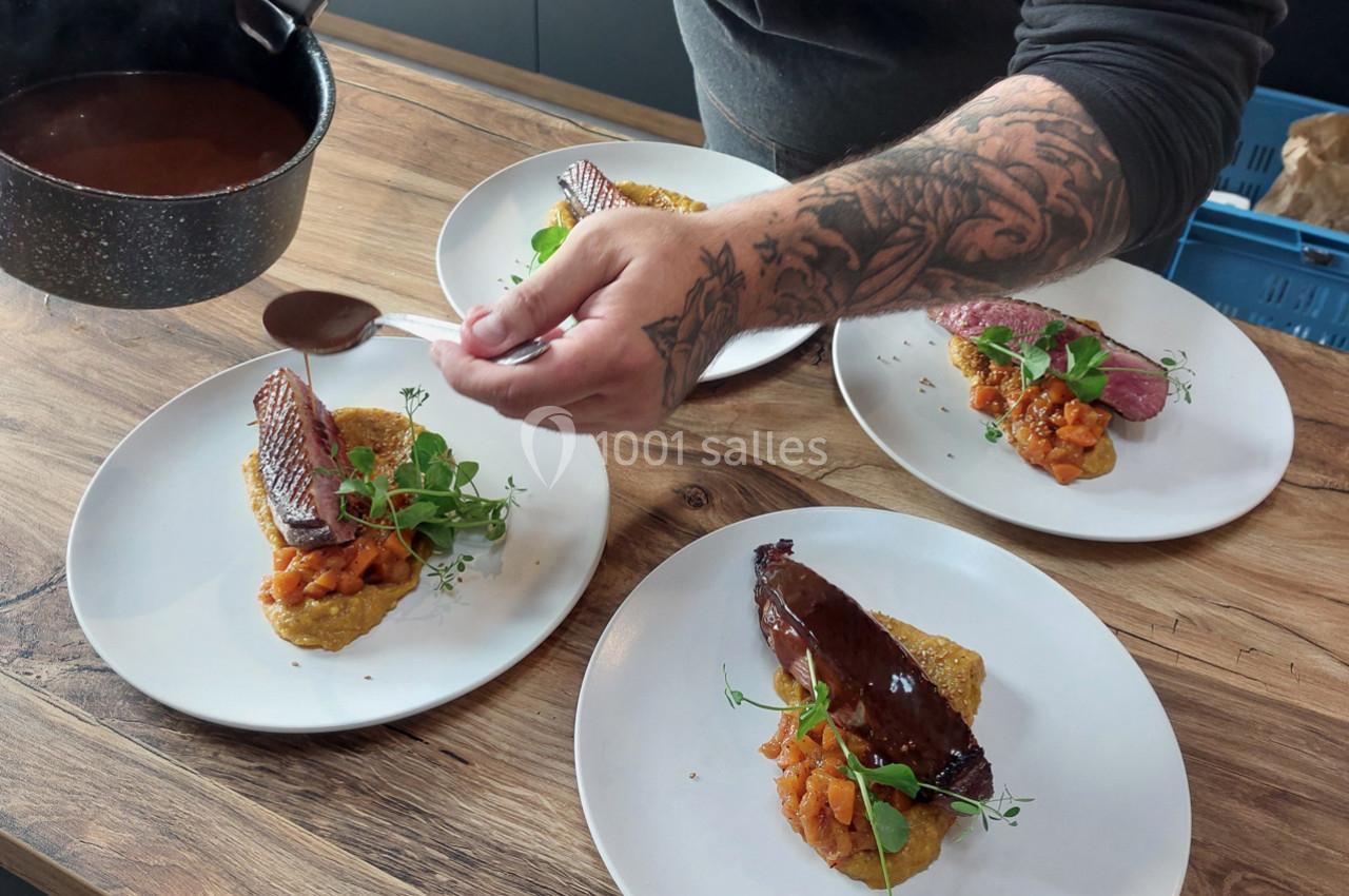 Un chef ajoute une sauce sur des assiettes garnies de viande, purée et légumes, disposées sur une table en bois.