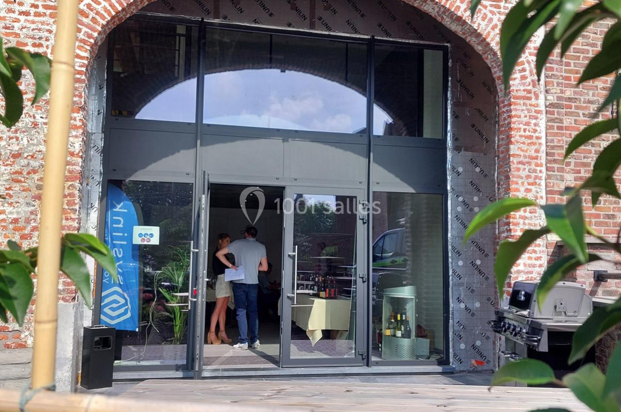 Entrée vitrée d'un bâtiment en briques, avec des personnes discutant à l'intérieur près de tables.