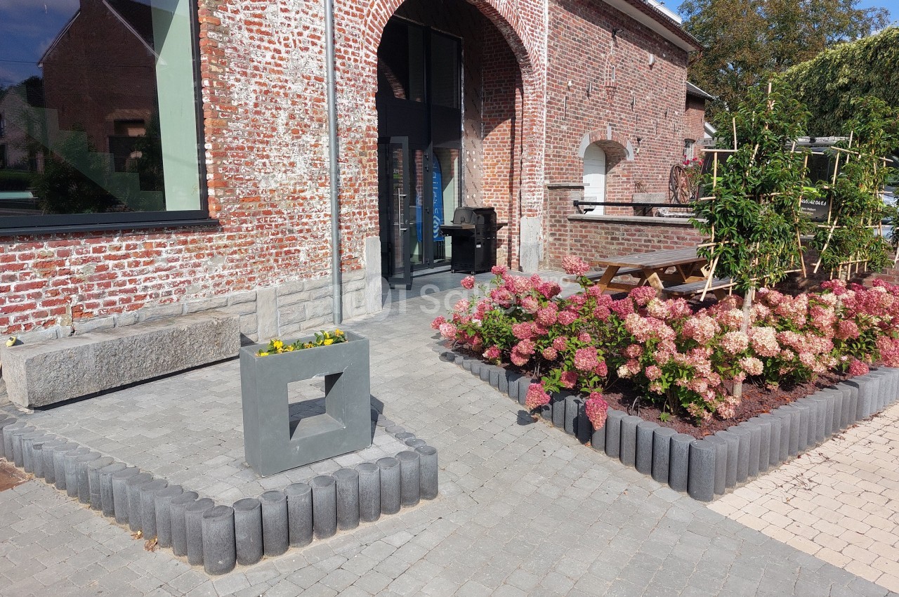 Entrée d'un bâtiment en briques rouges avec parterre de fleurs, pavés au sol et mobilier extérieur en bois.