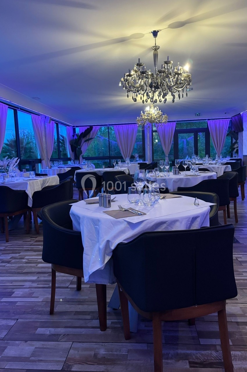 Salle de restaurant élégante avec tables dressées, chaises noires, nappes blanches et lustres imposants sous éclairage…