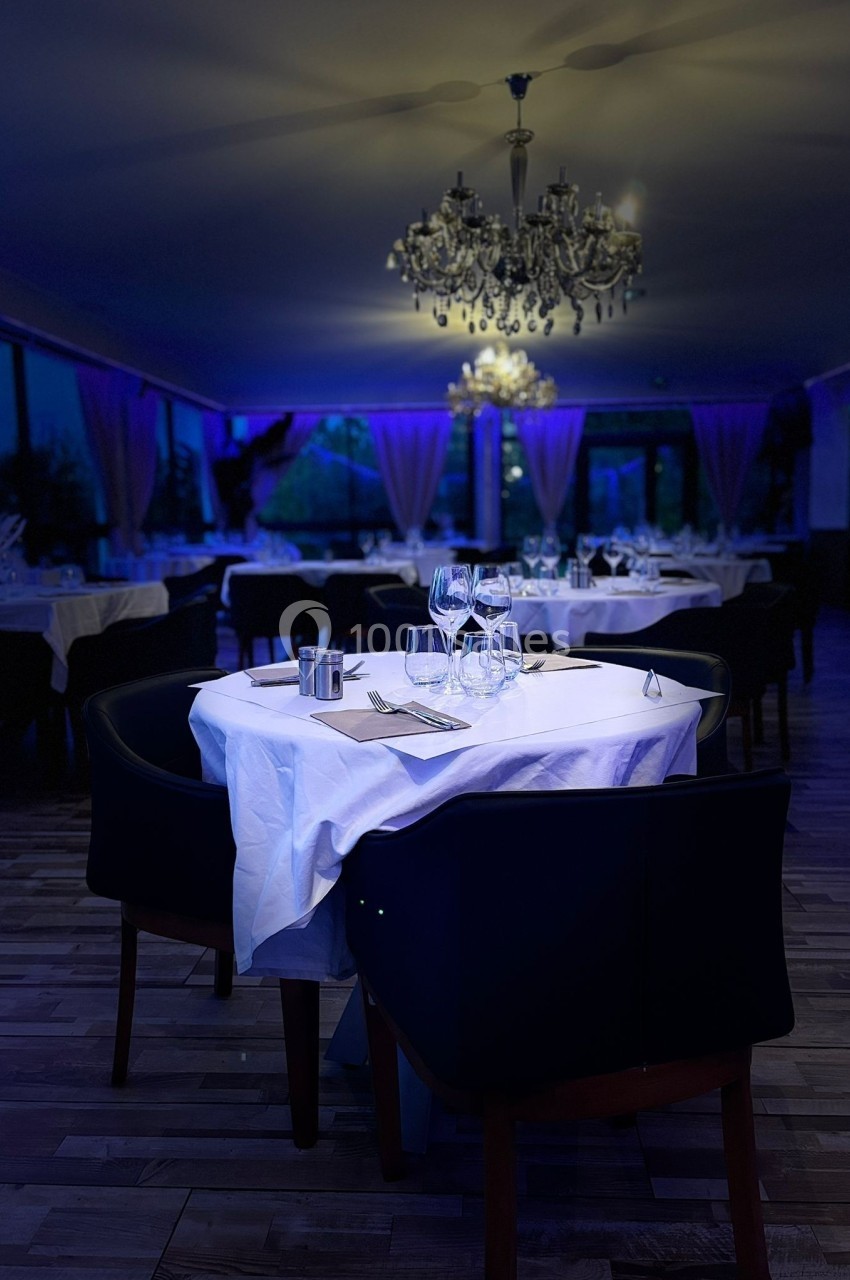 Salle de restaurant élégante avec tables dressées, nappes blanches, chandeliers et éclairage tamisé bleu.