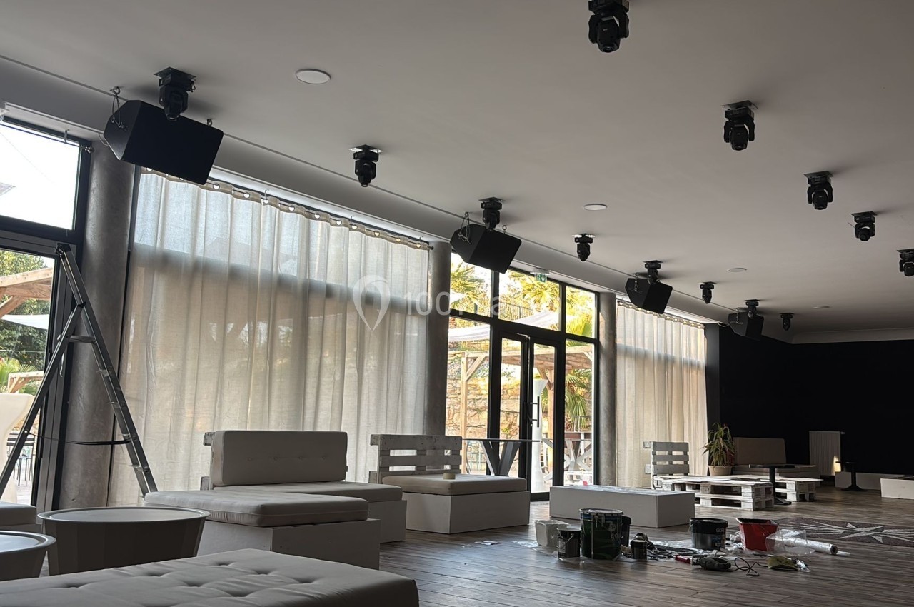 Salle lumineuse en cours d'aménagement, avec des canapés clairs, des rideaux transparents et des équipements au sol.