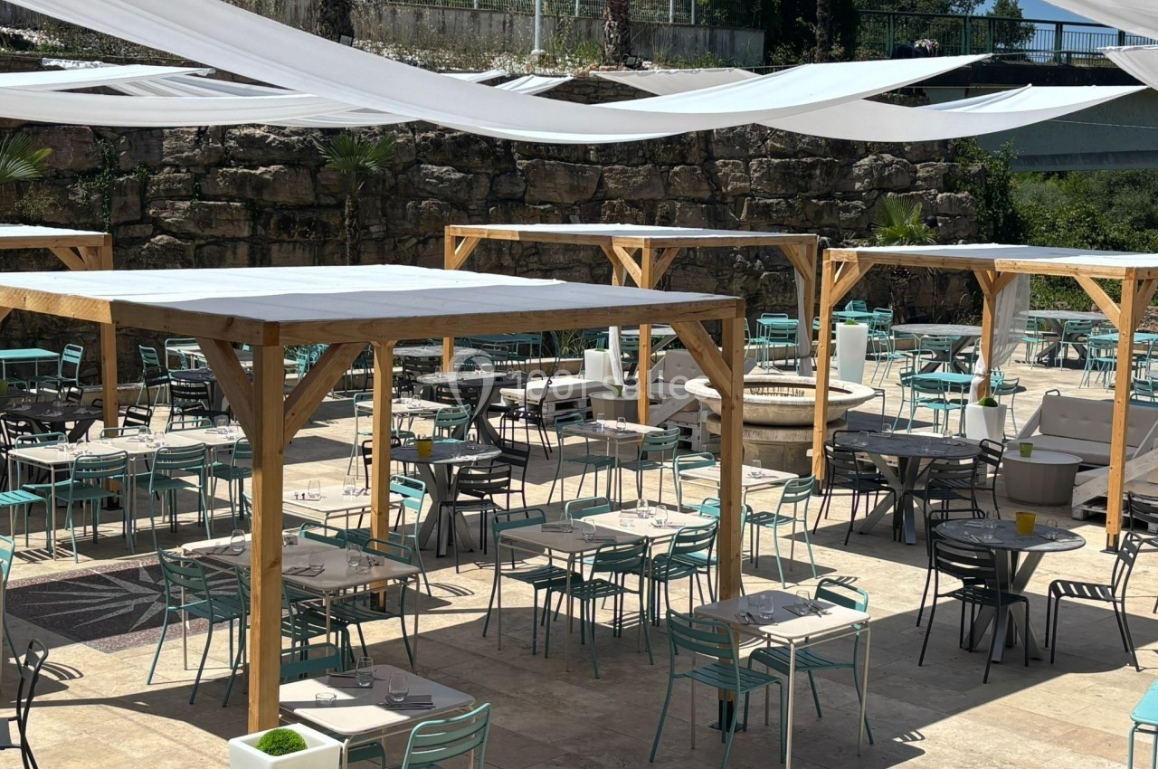 Terrasse extérieure avec tables et chaises sous des pergolas en bois, entourée de murs en pierre et de végétation.